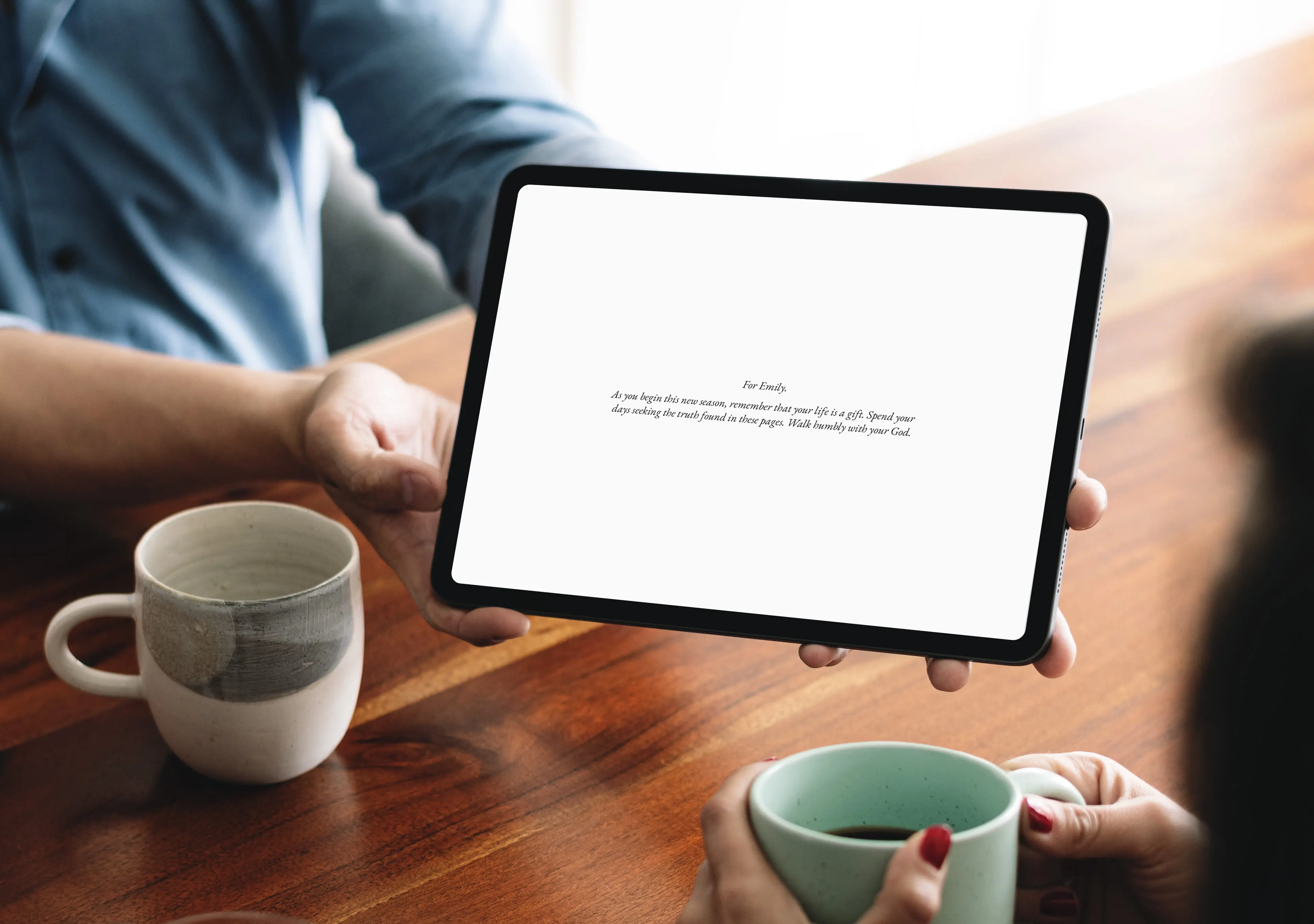 Person showing Personal Edition dedication page on tablet to another person, gift-giving moment over coffee