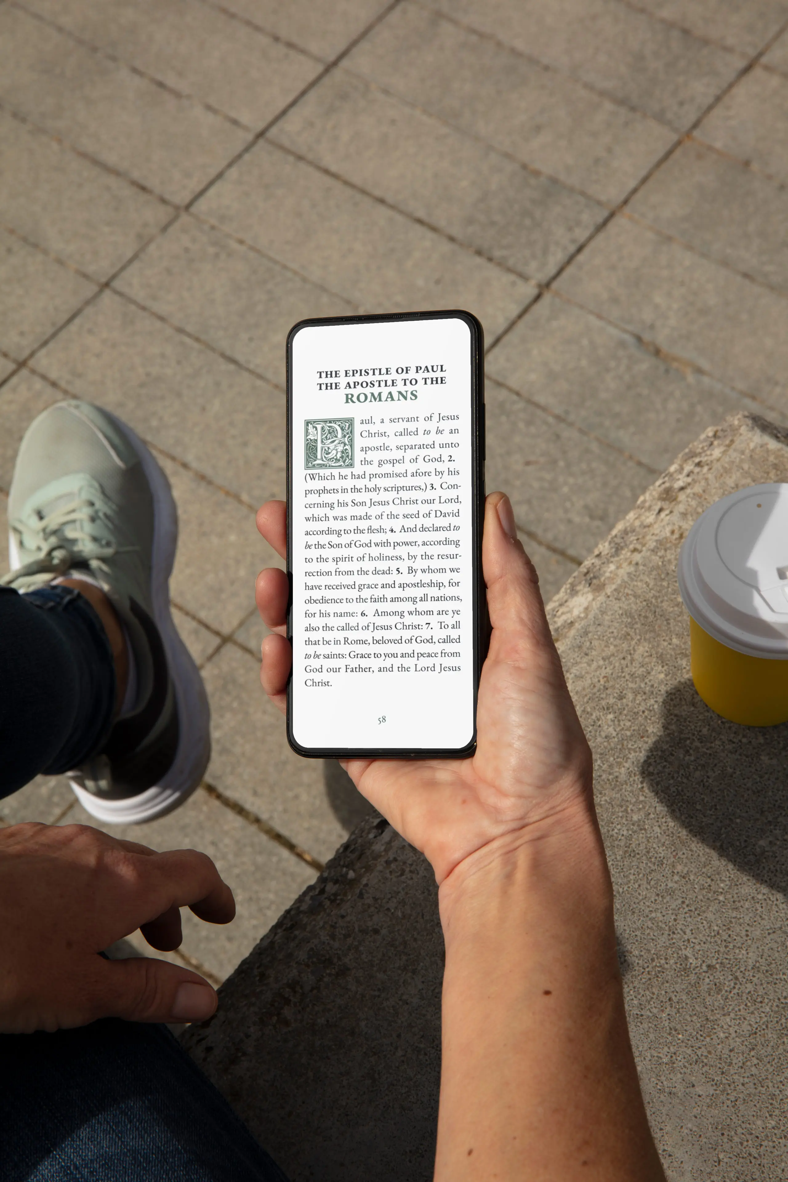 Hand holding smartphone displaying Romans from the large print KJV Bible while sitting outside with coffee