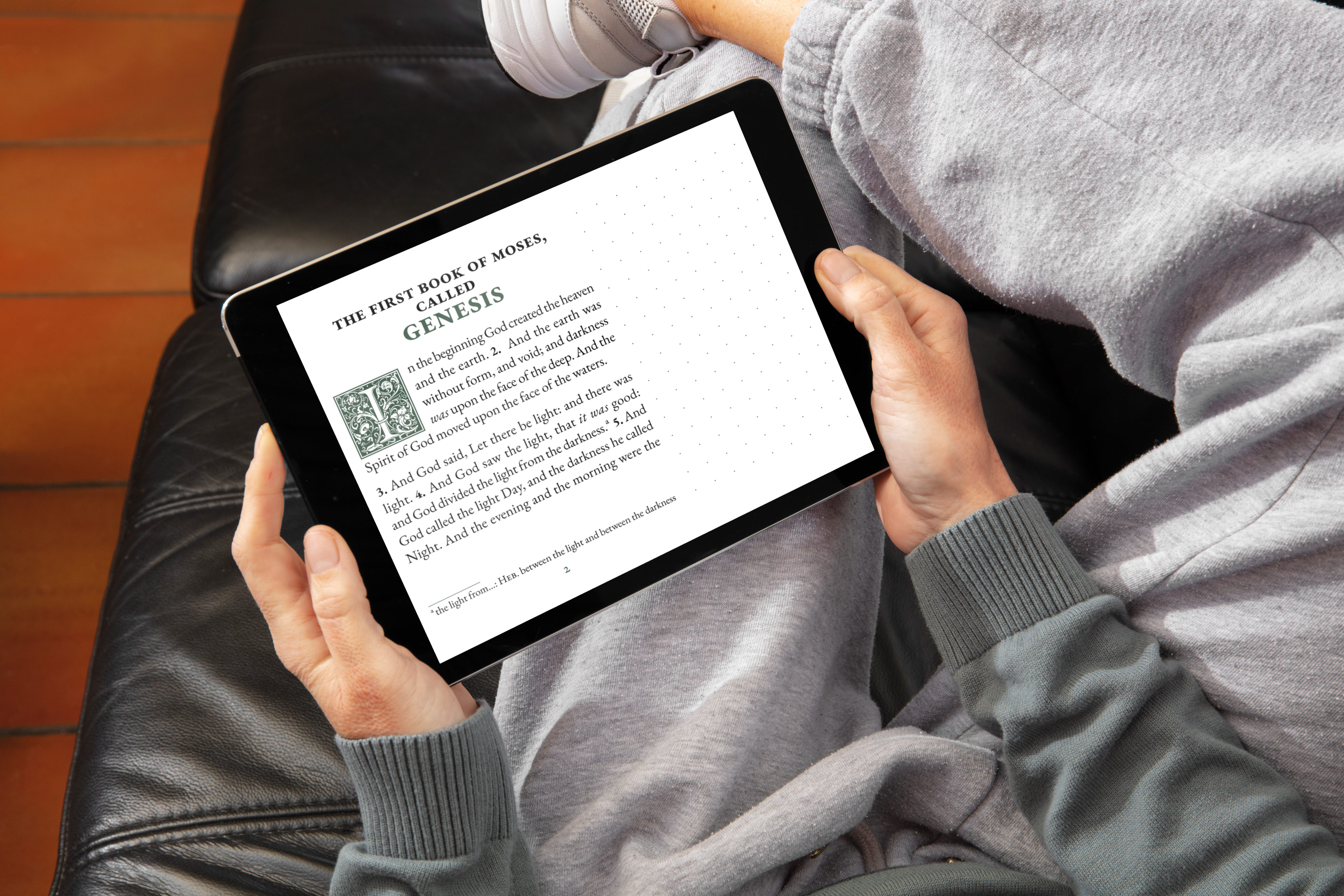 Person reading Genesis on tablet while relaxing on couch, showing large print text with dotted notetaking margin